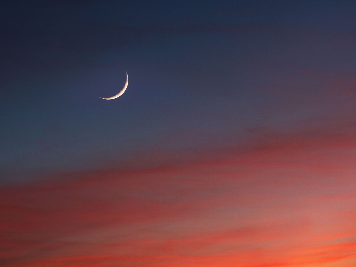 Schmale Mondsichel am Abendhimmel mit bunten Wolken in Orange und Blau