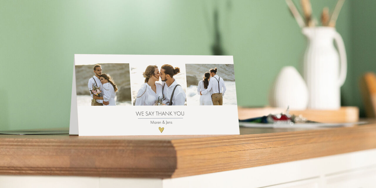 Une carte de remerciement au format paysage est posée sur une commode. Le recto est imprimé avec deux photos des mariés pendant le mariage sur un fond coloré. En dessous est imprimé « Merci beaucoup », ainsi qu’un clipart de trois cœurs.