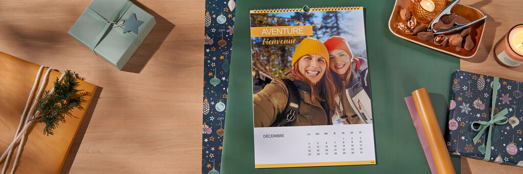 Un calendrier photo est posé sur une table entouré de feuilles de papier cadeau, d’un cadeau emballé, d’une bougie et d’un bol de noix. Sur la page du calendrier de décembre, on peut voir la photo de deux femmes souriantes vêtues de vêtements hivernaux ainsi que les mots « Aventure bienvenue ».