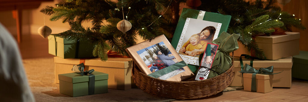 Un cesto regalo amorevolmente allestito con un fotolibro CEWE, un calendario e regali verdi sotto un albero di Natale
