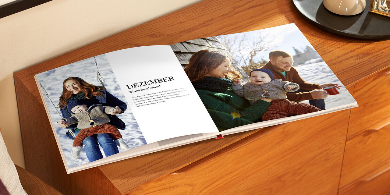Il fotolibro aperto è appoggiato su un mobile di legno. Sul lato sinistro c’è una foto di una donna e di un bambino e un blocco di testo con il titolo «Dicembre». Sul lato destro c’è una foto della famiglia per intero.