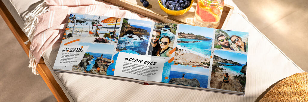 Un FOTOLIBRO CEWE aperto giace su un lettino con un telo da spiaggia con un vassoio con mirtilli e una bibita. Le pagine interne mostrano le foto di una coppia in vacanza nei toni del blu.